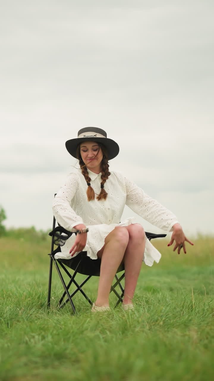 하 드레스와 검은 모자를 입은 행복한 여자가 잔디에 자리를 잡고 의자에 앉으려고 준비하고 있습니다. 색 셔츠를 입은 남자가 을 들고 예술적인 의도로 그녀를 관찰합니다.