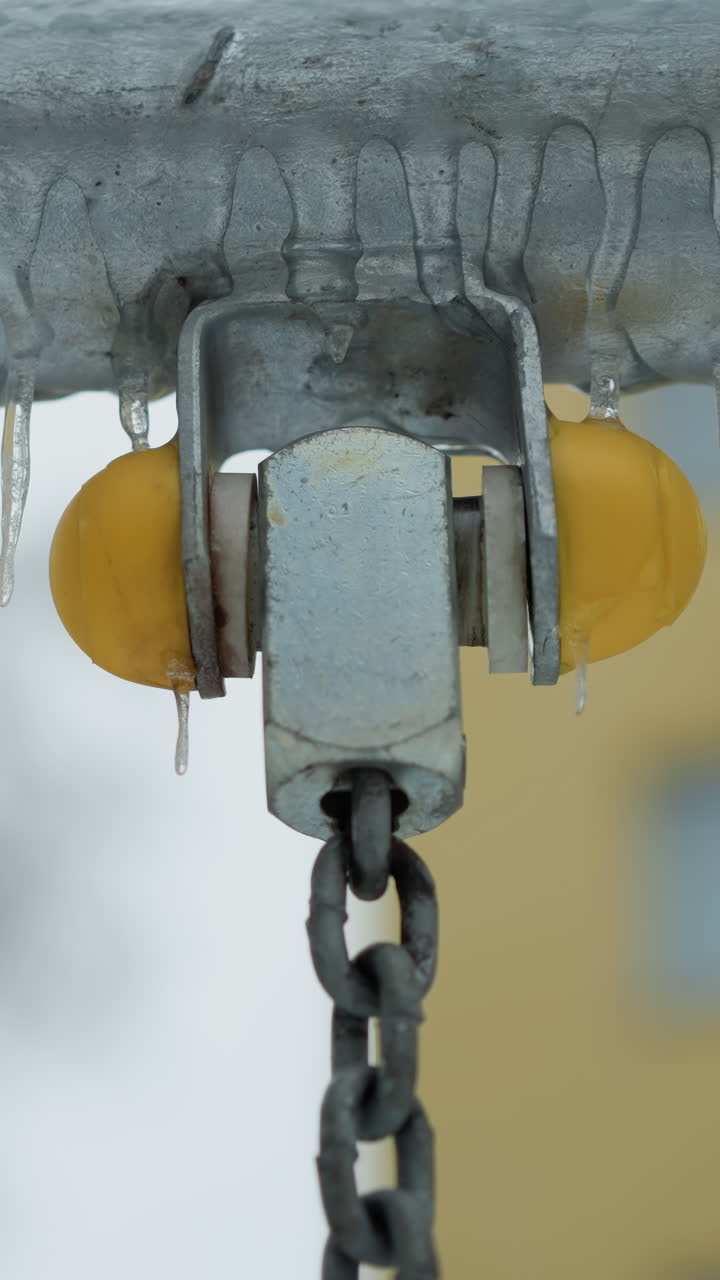 primer plano de una barra de metal con hielo colgado, mostrando detalles de juntas y cadenas de plástico amarillo, en contra de un fondo nevado borroso y un edificio amarillo débil
