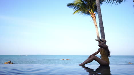 Hermosa-Mujer-En-Traje-De-Baño-Sentada-Al-Borde-De-Una-Piscina-Infinita,-Fondo-De-Vacaciones-De-Paisaje-Marino