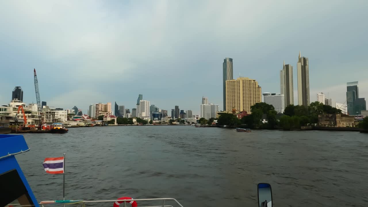 Câmera em primeira pessoa de um barco passeando pelo rio Chao Phraya, com os arranha-céus de Banguecoque ao fundo.