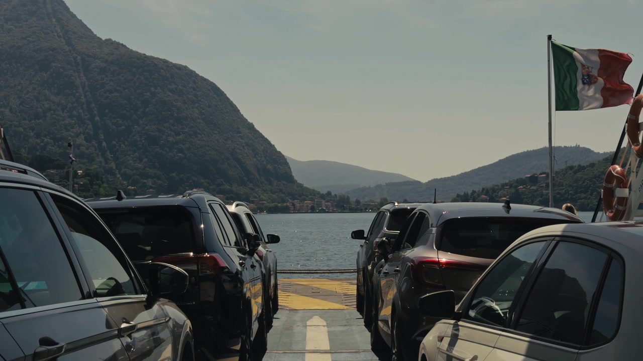 Vista en primera persona desde la cubierta de un ferry con coches aparcados a ambos lados, una bandera italiana ondeando al viento, aguas tranquilas del lago y un impresionante paisaje montañoso en un día soleado