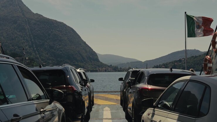 Vista en primera persona desde la cubierta de un ferry con coches aparcados a ambos lados, una bandera italiana ondeando al viento, aguas tranquilas del lago y un impresionante paisaje montañoso en un día soleado