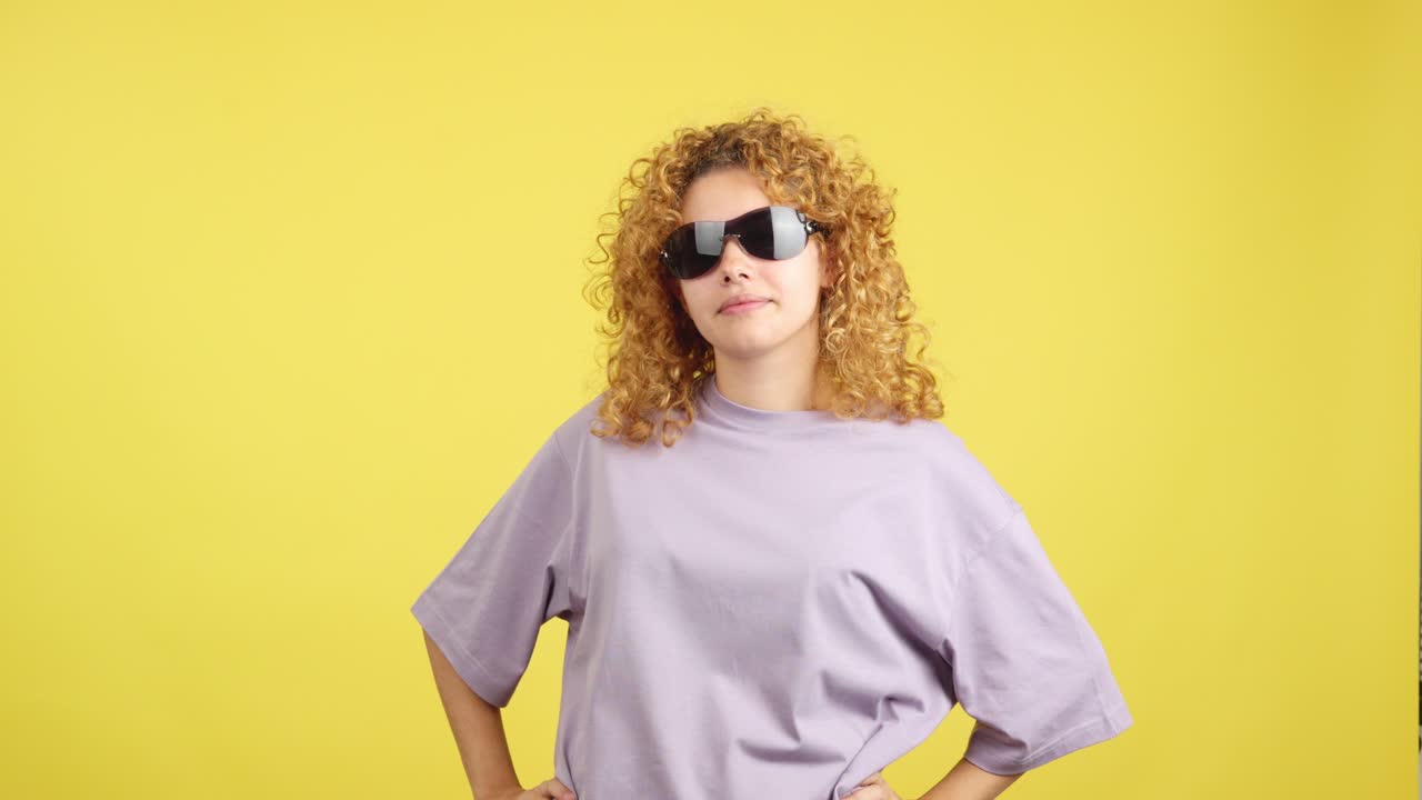 Confident young woman with curly blonde hair posing with sunglasses against a yellow background