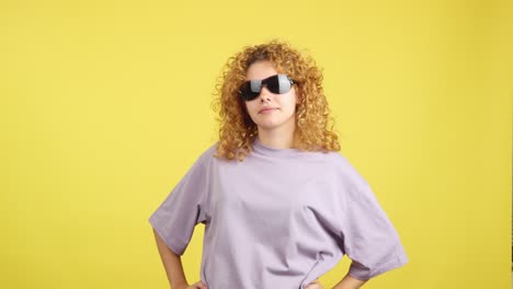 confident young woman with curly blonde hair posing with sunglasses against a yellow background