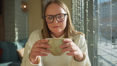 young woman in white sweater holding ceramic mug while sitting by large window decorated with string lights, bright winter sunlight shines through glass with snowy street visible in soft background