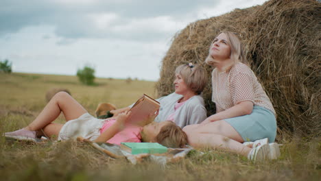 little girl in pink shirt reading her book with green cover, lying on picnic blanket, mother seated close adjusting her hair, another woman resting with dog on leash looking into distance