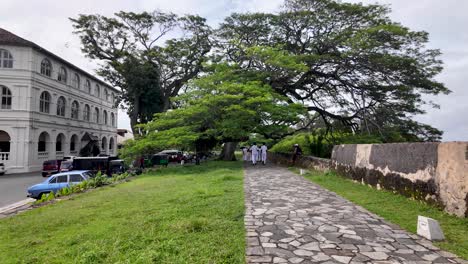 students in uniform stroll past a historic building and lush trees along a stone path in galle, sri lanka, capturing a blend of education and culture.