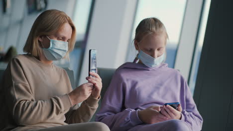 mother and daughter waiting for their flight in the airport terminal. wear protective masks, use smartphones