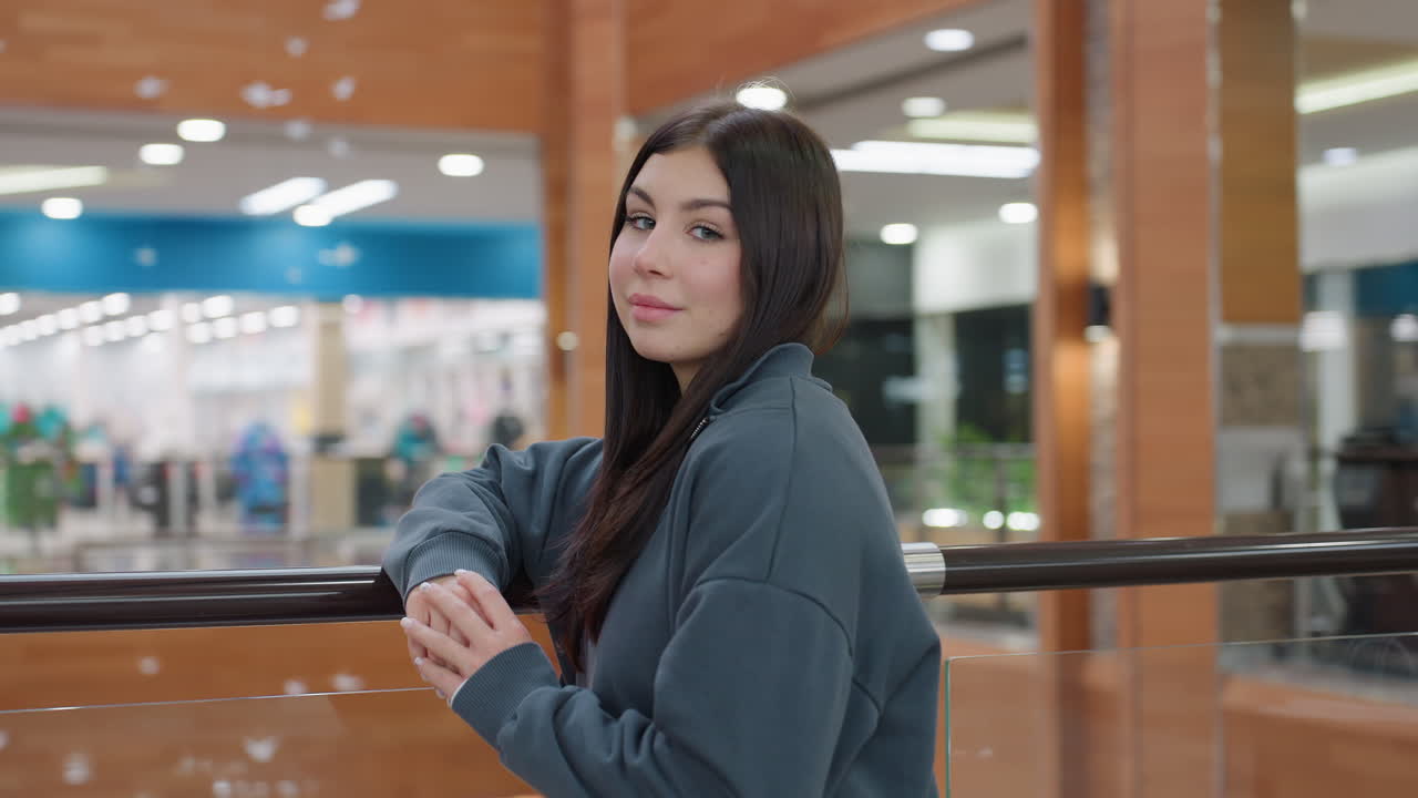 Vista frontale di una giovane e bellissima donna che appoggia le mani sulla ringhiera di un centro commerciale, con un sorriso gentile e un'espressione sicura, che indossa una felpa scura, in piedi all'interno di un moderno centro commerciale