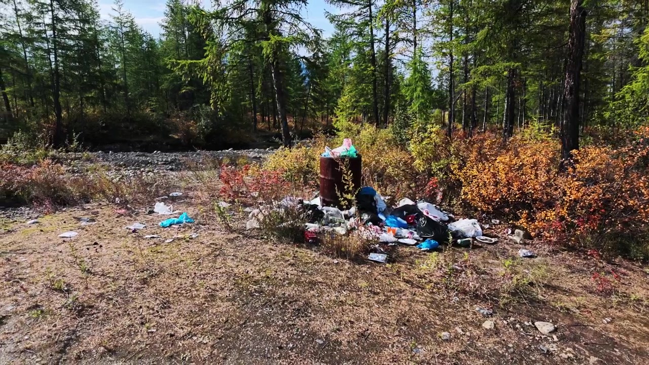 A slow zoom captures a pile of plastic bags, bottles, and debris scattered around a rusted barrel in a forest clearing near a stream, surrounded by trees and autumn-colored bushes.