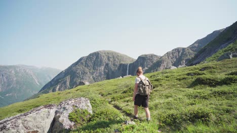 Young-Man-Walking-On-Meadows-At-The-Mountains-With-Mardalsfossen-Waterfall-In-Norway