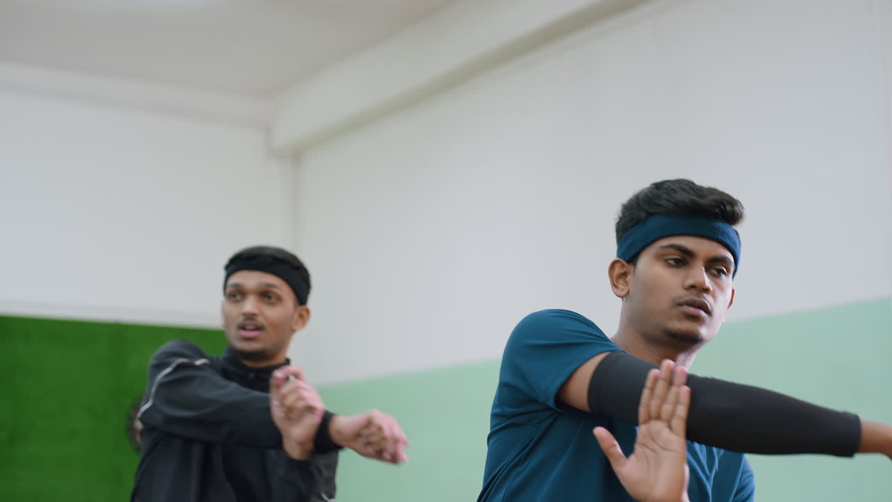 Two friends wearing sports headbands perform arm stretching exercises during indoor workout session, focusing on flexibility, coordination, and preparation for athletic performance in training hall