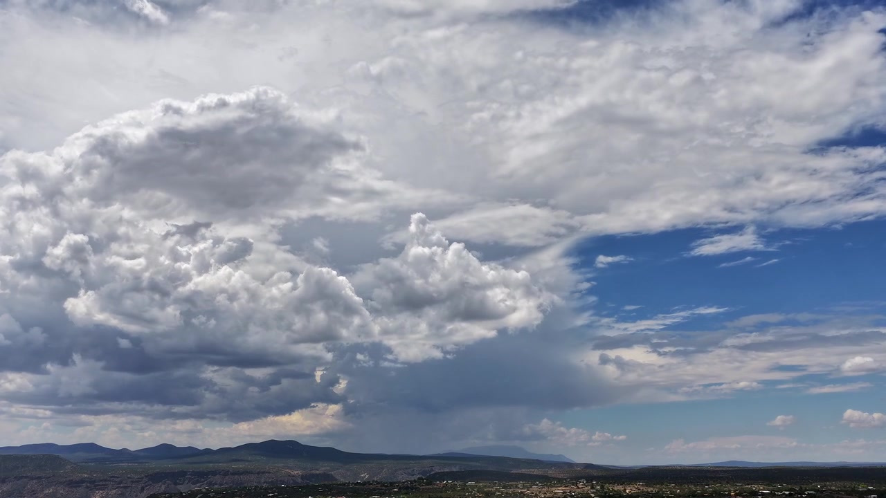4K drone timelapse observing a massive, multi-layered storm system gathering over Los Alamos County, New Mexico. Towering white and grey cumulonimbus clouds dominate the sky above the rugged mountains