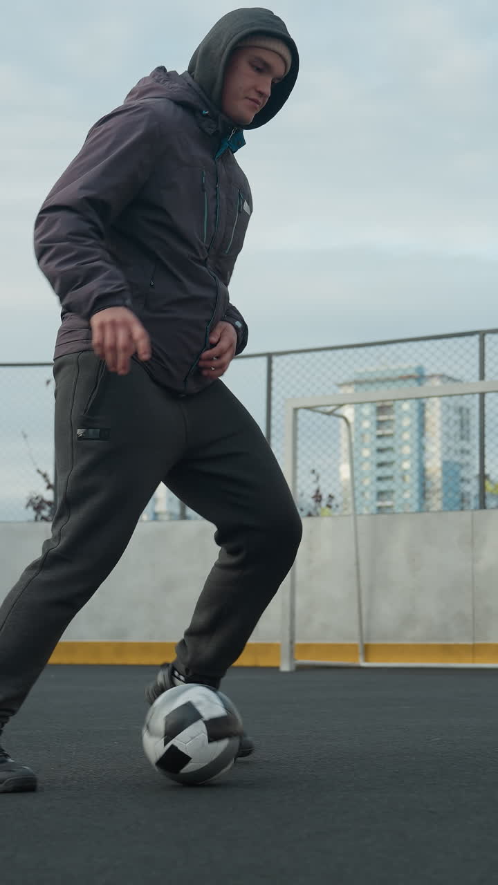 Young man skillfully controlling soccer ball with precise footwork on sport arena, showcasing balance, agility, and athleticism, background includes urban architecture and blurred goal