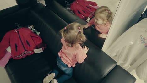 una niña pequeña en una tienda de ropa se sienta en el sofá frente a un espejo