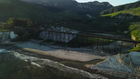 Aerial-orbit-descends-to-rocky-cliffs-with-railroad-trestles-and-bridge-on-sand-at-Gaviota-Beach,-California-at-sunrise