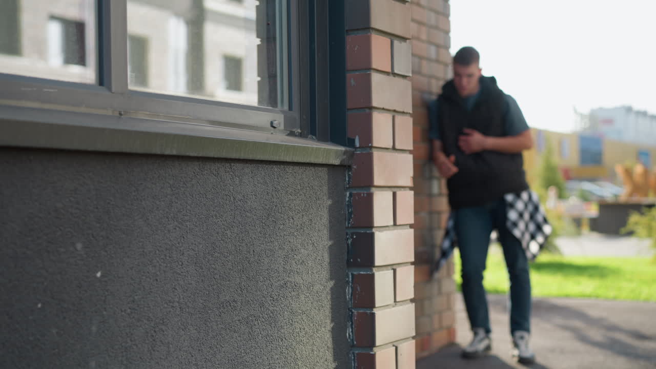 blurred view of young male student in casual outfit leaning on brick building corner while inhaling from vape device and coughing on sunny day with urban street and building background