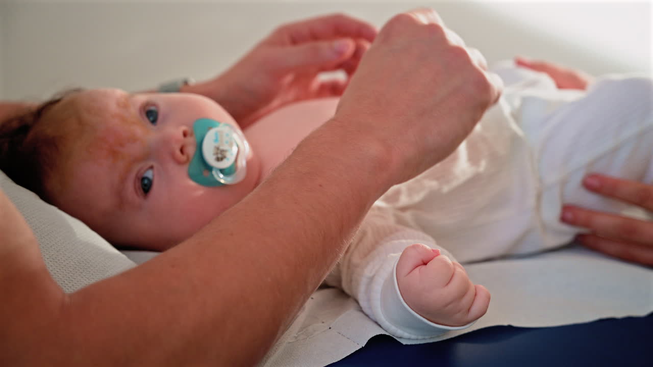 Close up of a baby lying calmly on a medical examination table, holding a pacifier while an adult gently supports them