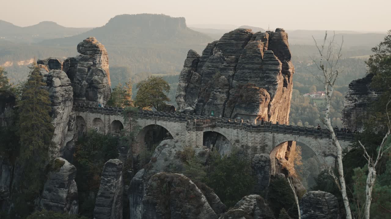 прекрасный вид на знаменитом бастионном мосту в саксонии, германия, недалеко от дрездена во время настроенного захода солнца, в то время как люди проходят по нему, чтобы исследовать место и иметь потрясающий вид на пейзаж с камнями