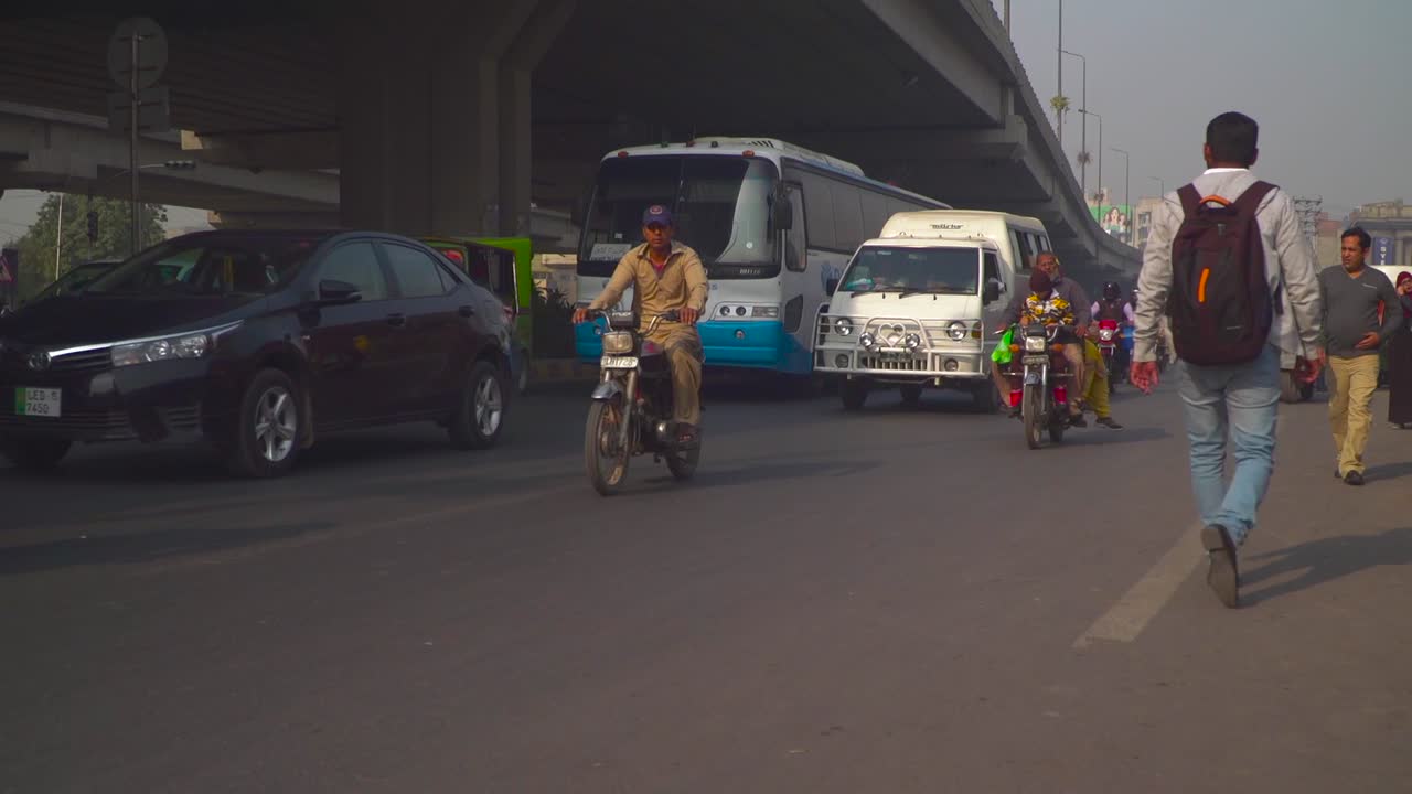 Traffic close up at the road under the fly over, passing motor bikes and cars, cycle, Buses, rickshaw`s, Billboards and buildings in the background