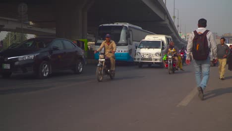 traffic close up at the road under the fly over, passing motor bikes and cars, cycle, buses, rickshaw`s, billboards and buildings in the background