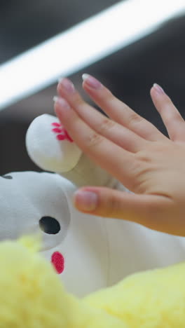 dama de rosa toca suavemente la mano de un juguete de peluche blanco en un estante en una tienda bien iluminada, ella toca la tela suave con cuidado, con otros juguetes alrededor de la tienda, con la tienda bien iluminado