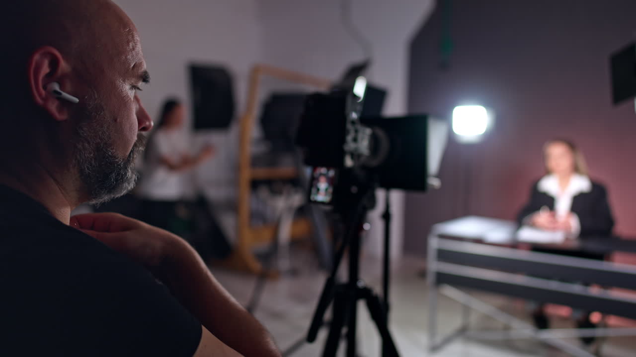 Bearded photographer looks focused on camera display. Cameraman filming a lady reporter sitting at desk at blurred backdrop. Studio backstage.