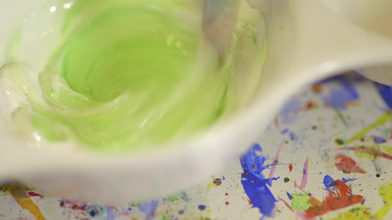Close-up de um artista misturando tinta verde e branca em uma paleta de cerâmica com um pincel. Processo criativo mostrando mistura de cores, textura e preparação artística.