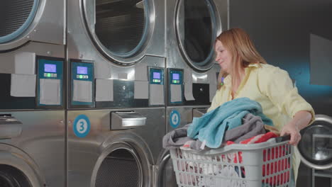 female worker carries basket of clothes, walks away from industrial washer inside laundromat, machines in row, piles of towels visible, routine service, clean facility, daily chores, smooth operation