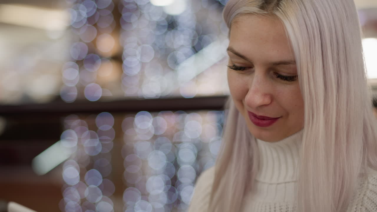 portrait of white lady seated on bench in bright mall wearing cozy sweater tapping smartphone with gentle smile and occasional hair adjustment under soft bokeh lights near glass railing