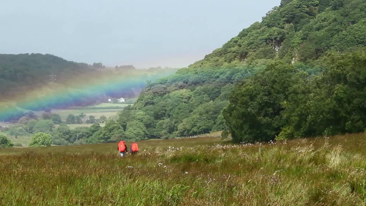Two hikers trekking through a green landscape with a rainbow above them