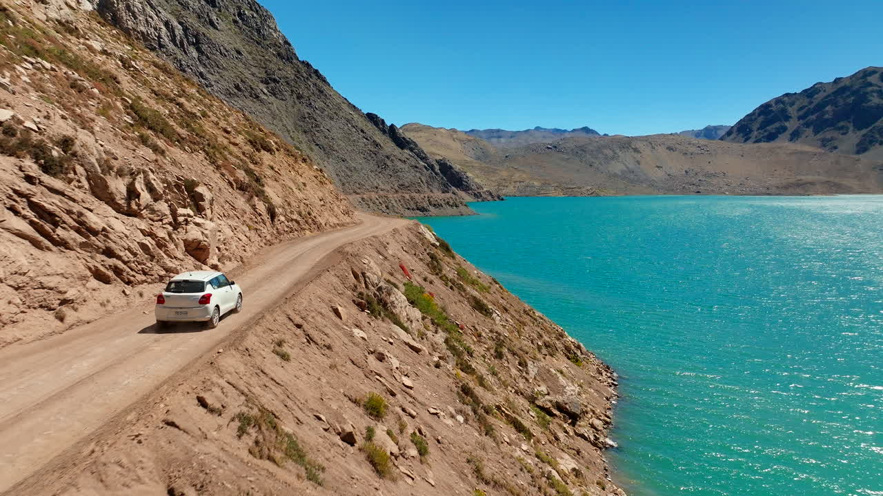 Aerial drone view of car driving on dirt road alongside turquoise Embalse El Yeso reservoir in Cajon del Maipo, Andes mountains, Chile