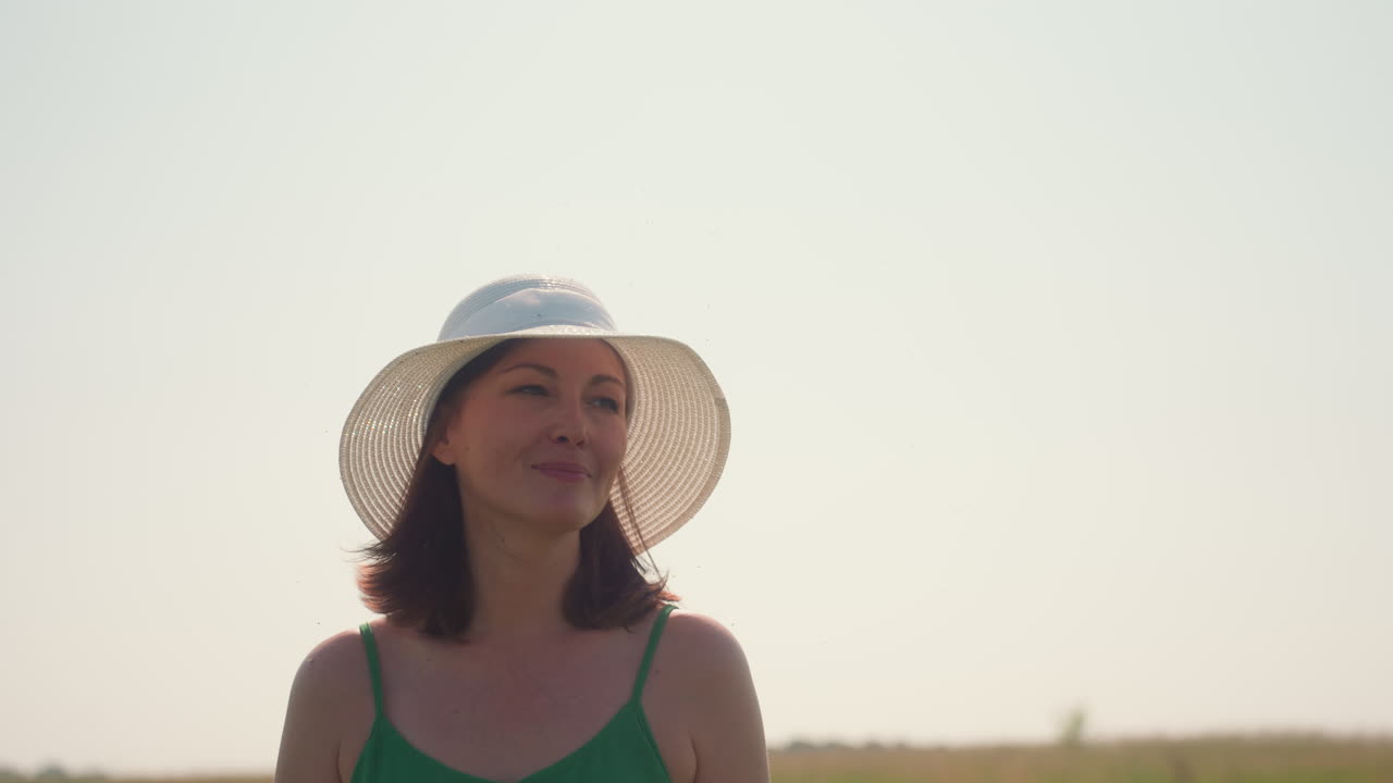 Closeup of smiling woman in green dress and white sunhat walking outdoors under clear sky, enjoying sunlight and summer breeze in peaceful countryside with natural warmth