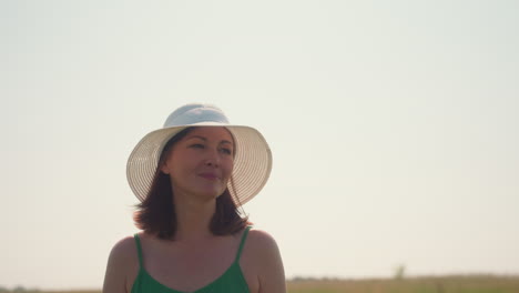 closeup of smiling woman in green dress and white sunhat walking outdoors under clear sky, enjoying sunlight and summer breeze in peaceful countryside with natural warmth
