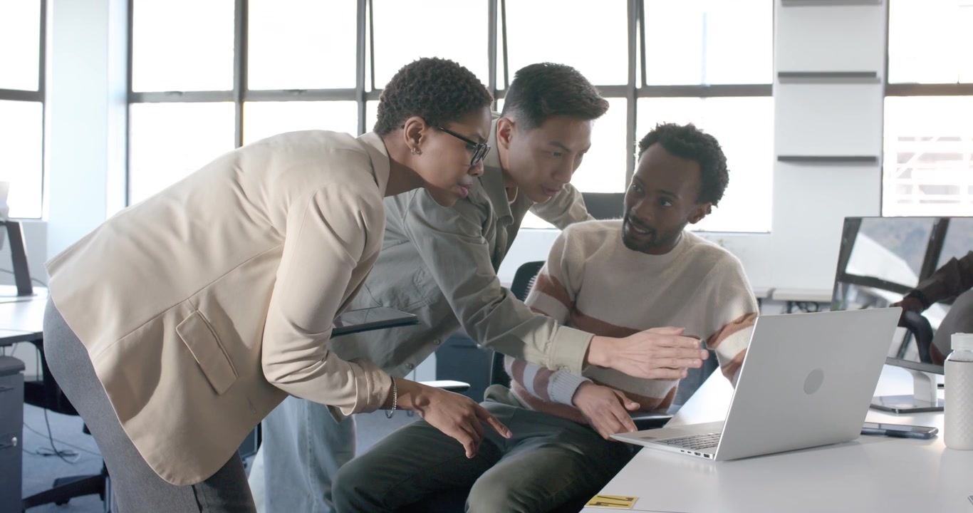 Diverse coworkers clustering around laptop at desk, being guided by arriving colleague with tablet