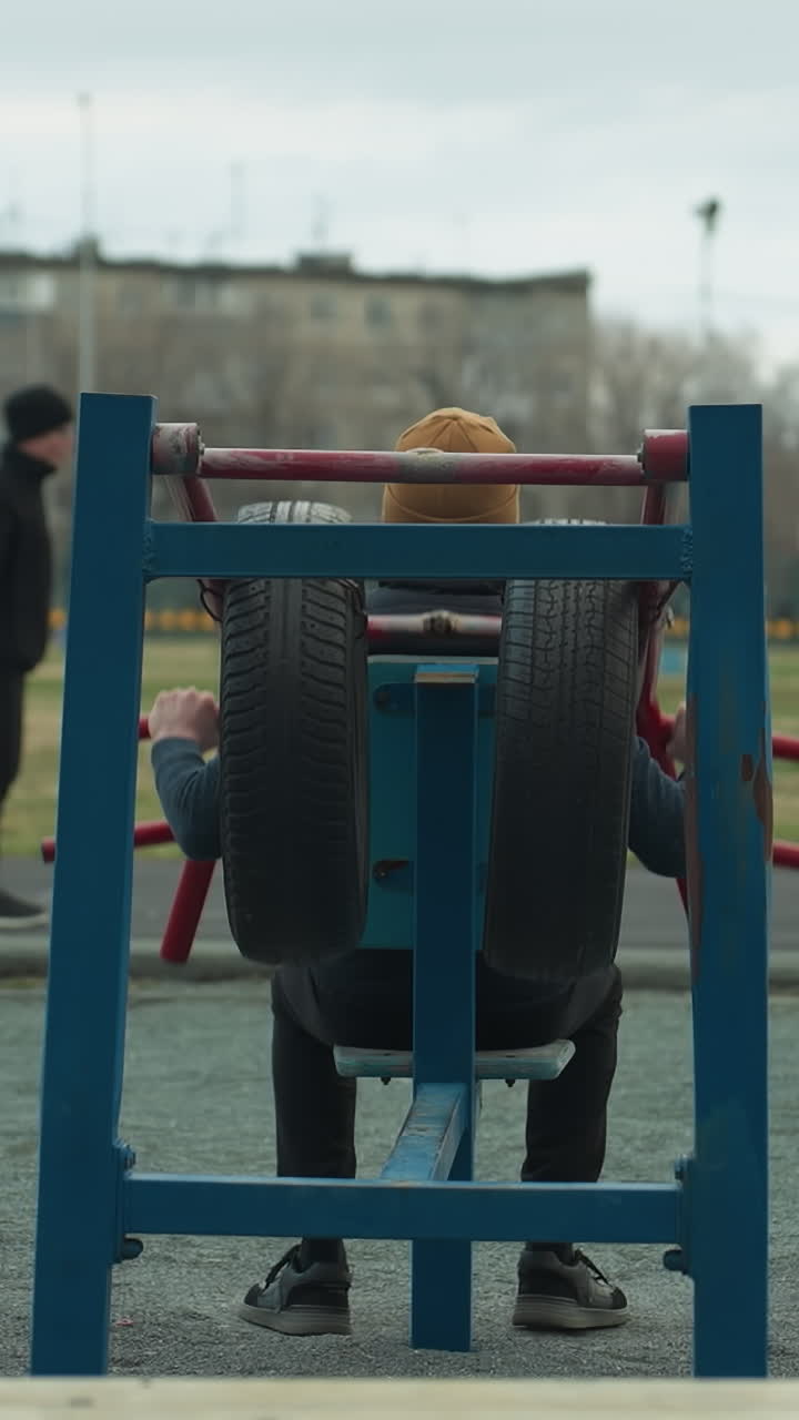 Close-up of a coach gripping workout equipment while exercising outdoors,two boys are also working out on a track near a grassy field, with people visible in the background