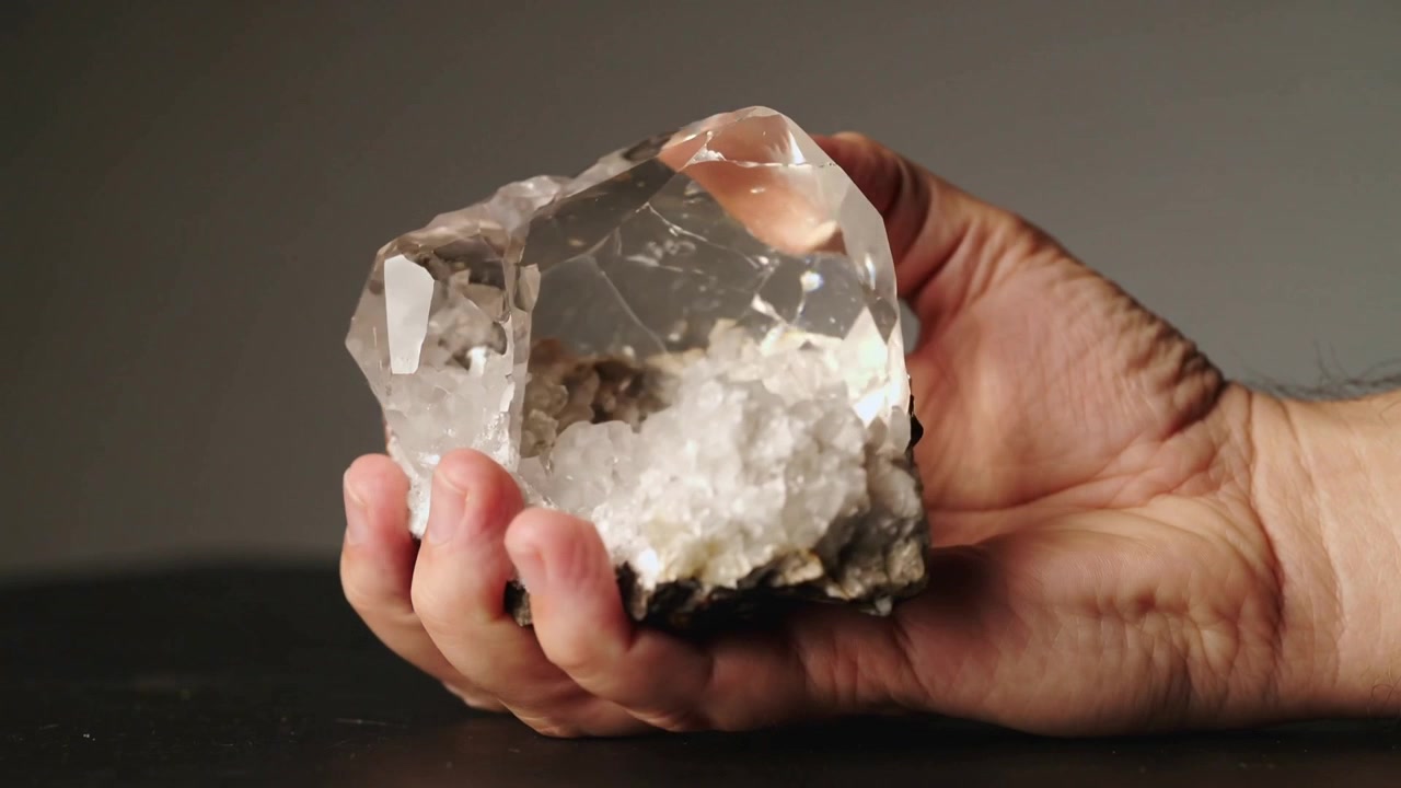 Grasping right hand, male holder rotating clear quartz cluster on dark tabletop, revealing druzy