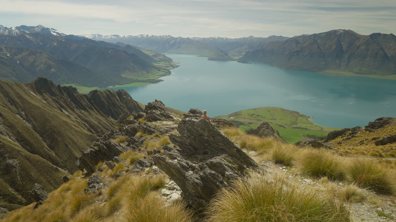 excursionista rubia corriendo hacia el borde del acantilado con impresionantes vistas del lago hawea