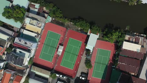 Aerial-top-down-rotation-of-red-and-green-tennis-courts-in-a-clockwise-direction-featuring-small-canal-and-rooftops-of-surrounding-buildings