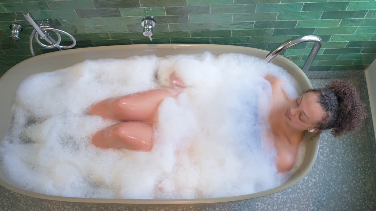 Una donna è sdraiata nella vasca da bagno con abbondante schiuma, nota una bottiglia verde e si allunga per ispezionarla.