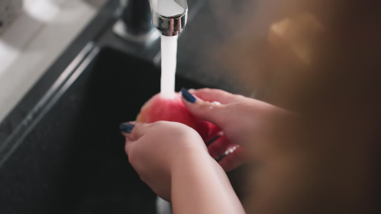 Overhead view teenager hands washing red apple under running water at kitchen sink, blue nails, thorough rinse, splashes and droplets, hygiene first, clean produce prep for healthy snack