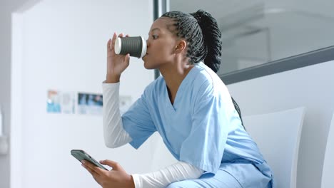 african american female doctor using smartphone, drinking coffee in corridor, slow motion