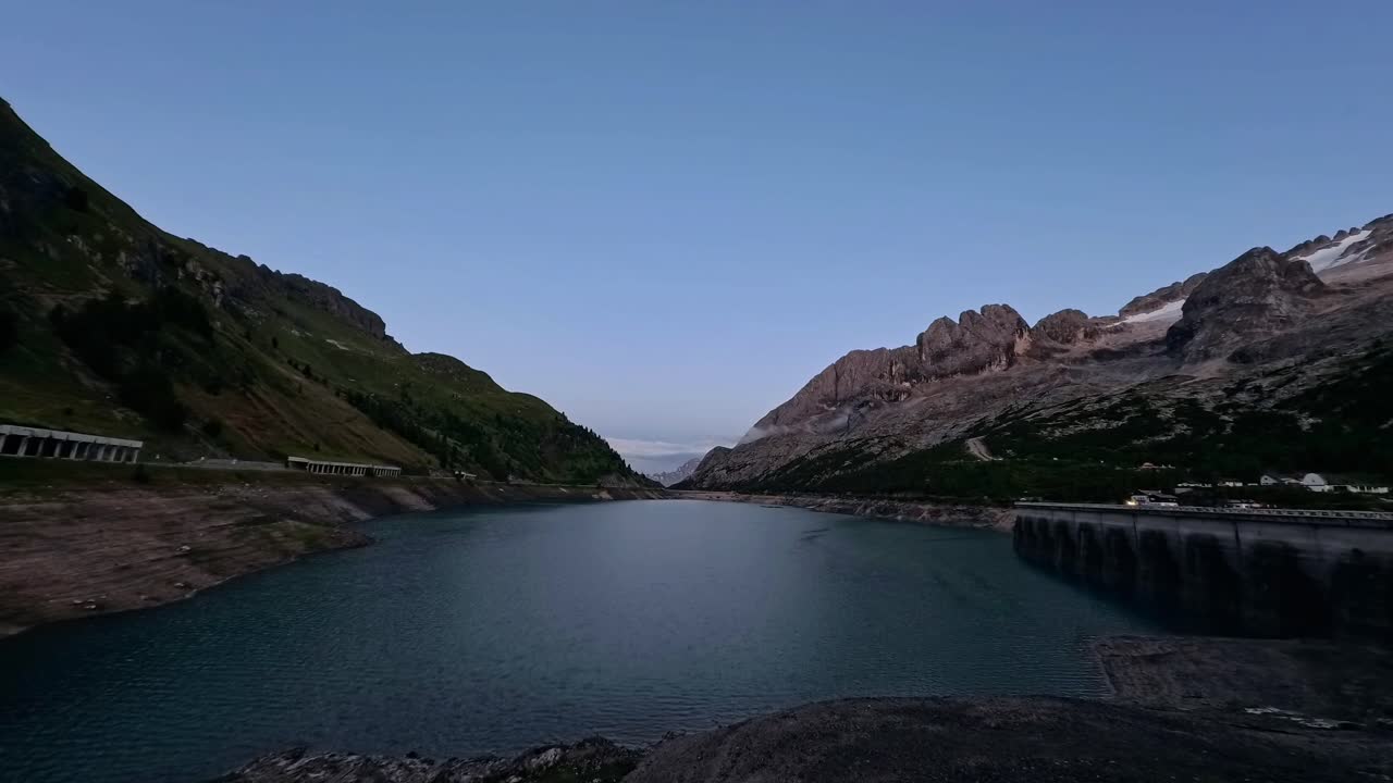 Il lago di Fedaia in Italia all'ora del crepuscolo con montagne, nuvole e acqua blu. Il paesaggio pittoresco include una diga e delle verdi colline, che lo rendono perfetto per i viaggi