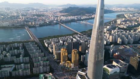 Timelapse-Drone-Shot-De-Lotte-World-Tower-Al-Atardecer-Con-El-Distrito-De-Songpa-Y-El-Río-Han,-Seúl,-Corea-Del-Sur