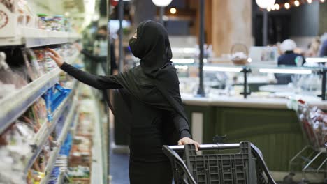 Woman-in-hijab-and-protective-mask-doing-shopping,-takes-product-from-the-shelf