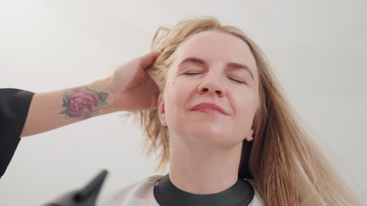 Una donna caucasica si fa fare una piega in un salone e sorride mentre la stilista con l'asciugacapelli e la mano tatuata crea una brezza calda, asciugando e arruffando delicatamente i lunghi capelli biondi; la cliente chiude gli occhi e si appoggia all'indietro sotto la luce intensa