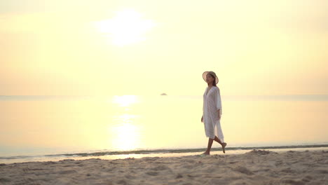 Mujer-Asiática-Caminando-En-La-Playa-Junto-Al-Agua-Del-Mar-En-Una-Brillante-Puesta-De-Sol-En-Verano-Usando-Un-Vestido-Blanco-Y-Un-Sombrero-De-Paja---Tiro-Estático-En-Cámara-Lenta