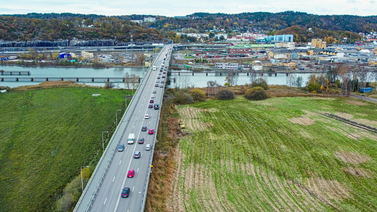 Vista aerea con drone del ponte Jordfallsbron vicino a Kungalv, che trasporta traffico attraverso il fiume Nordre Alv tra la terraferma e Hisingen, con terreni agricoli circostanti, corsi d'acqua e paesaggio urbano.