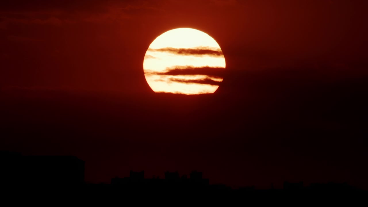 Large bright sun centered in a deep red sky above a distant urban skyline