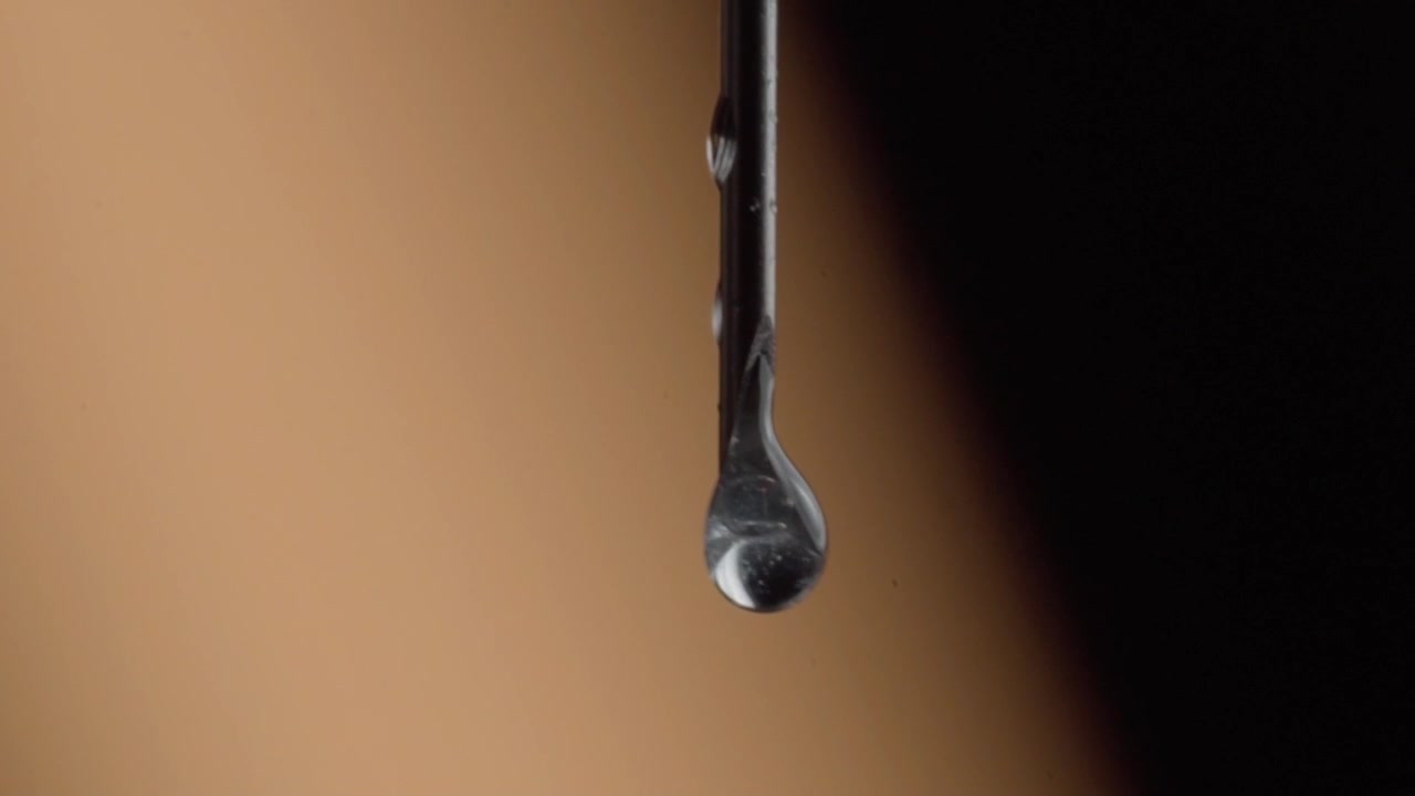 Macro close up of a liquid droplet suspended from the tip of a glass dropper, capturing surface tension moments before the drop falls